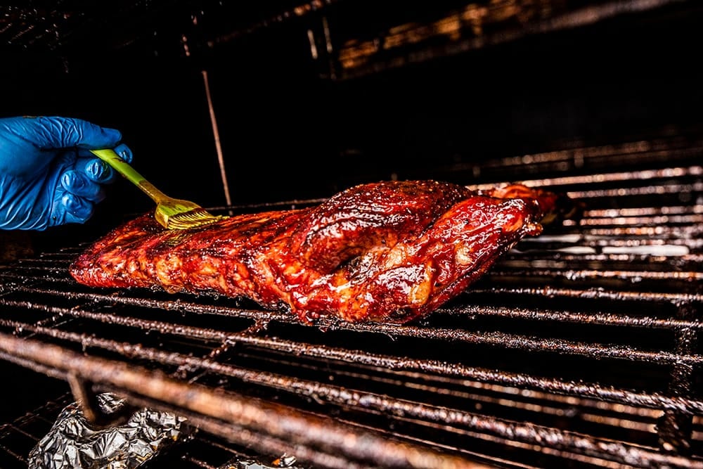 A gloved hand basting sauce onto racks of ribs laid across the grill bars of the cooker.