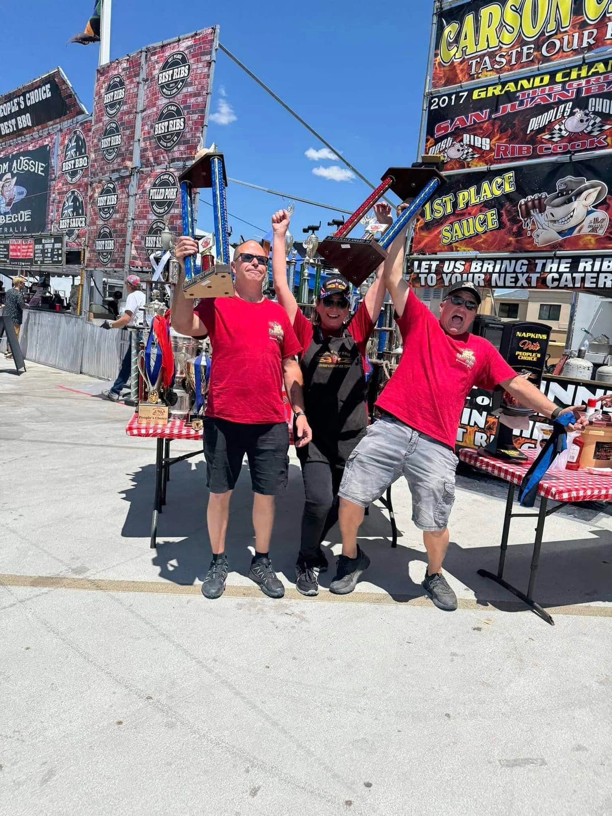 Three men in red Carson City BBQ shirts jumping and cheering in front of the team's competition booth, whose banners read "1st Place Sauce" and "Let us bring the ribs to your next cater".