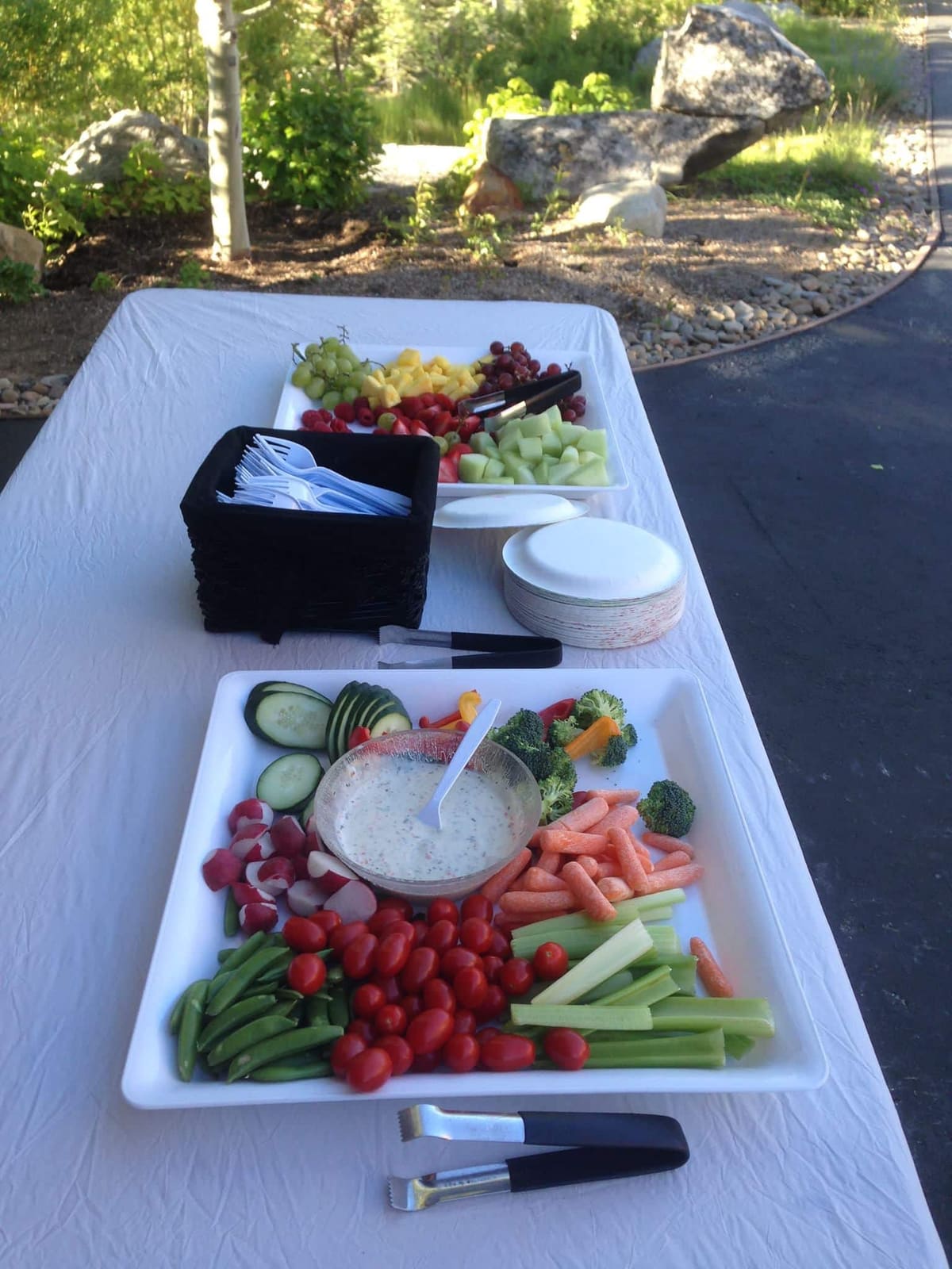 A white platter of raw vegetables, grapes and dip laid out as an appetizer tray on a white-clothed table outdoors.