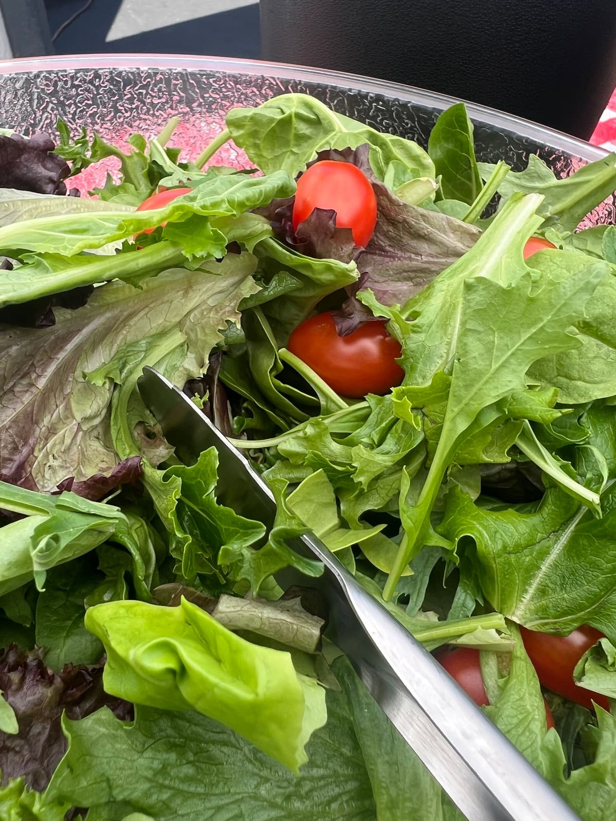 A tossed green salad of mixed leaves and cherry tomatoes being served with tongs from a large bowl.
