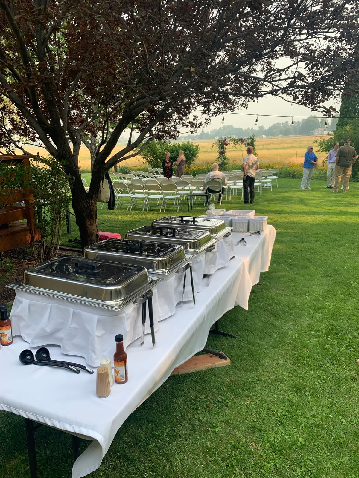 Chafing dishes and a bottle of sauce along a white-clothed buffet table on a lawn beneath a tree, with a ceremony seating area set up in the background.