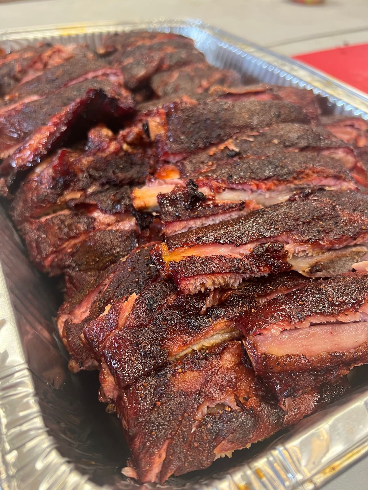 A full rack of dark, bark-crusted ribs resting in a foil tray, ready to be portioned.