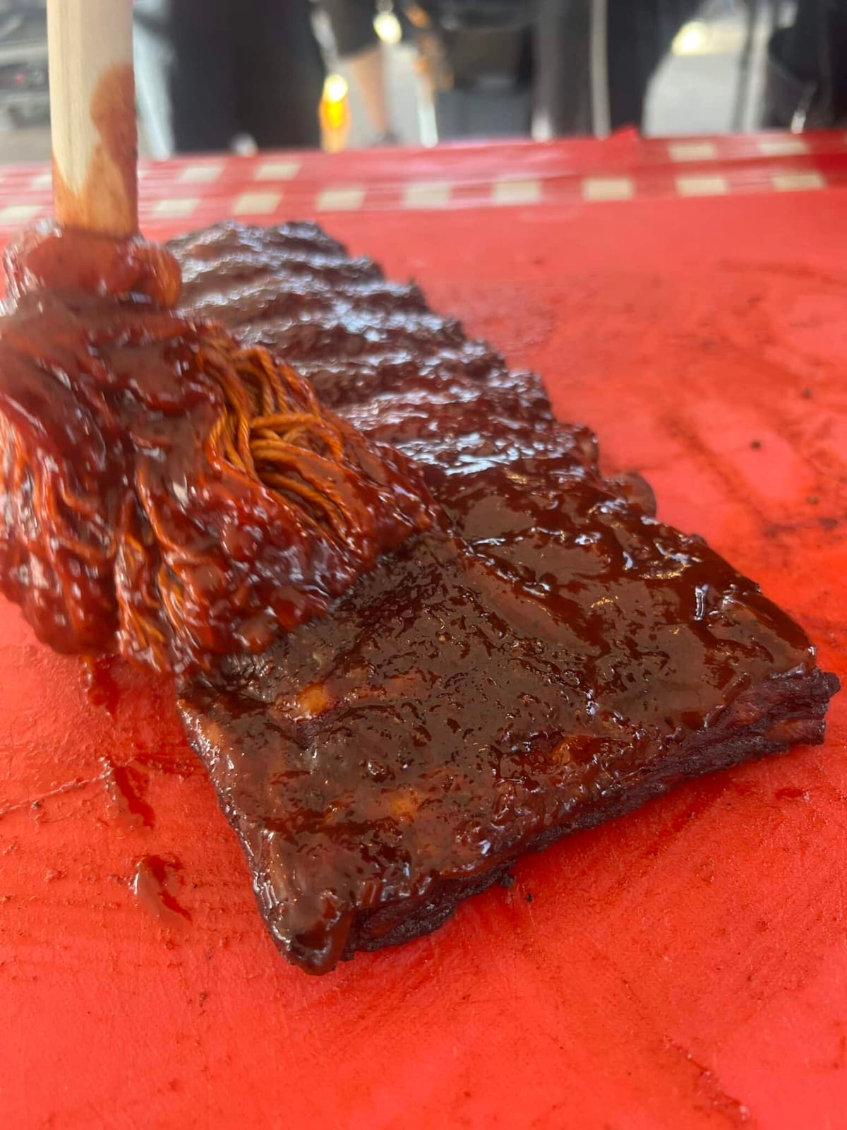 A basting brush drawing a stripe of sauce down a rack of ribs on a red cutting board.