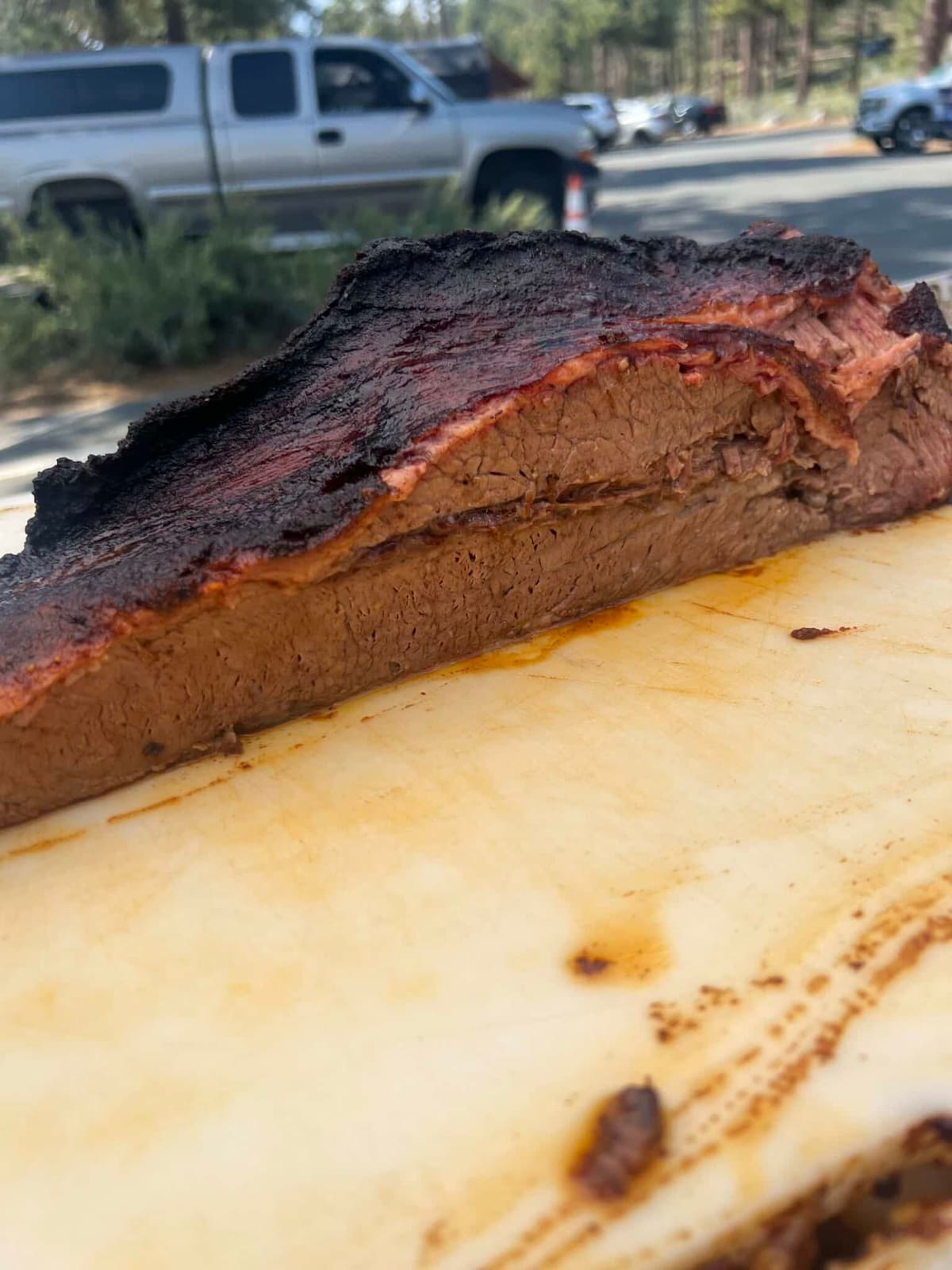 A close-up of a smoked brisket point resting on a board, with a pickup truck and roadway blurred behind it.