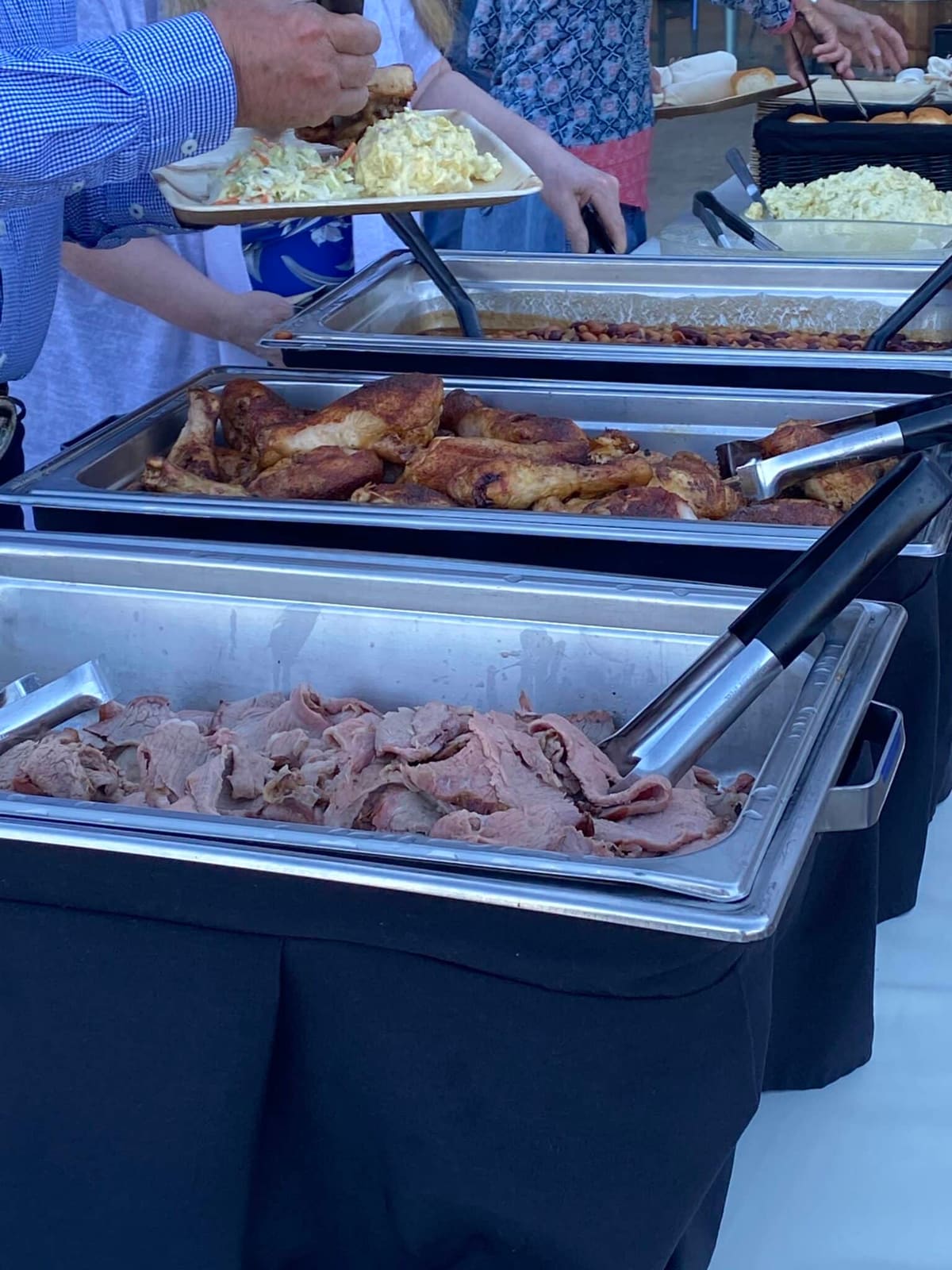 Chafing trays of sliced meats and a hand lowering a tray of coleslaw into place along an outdoor buffet line.