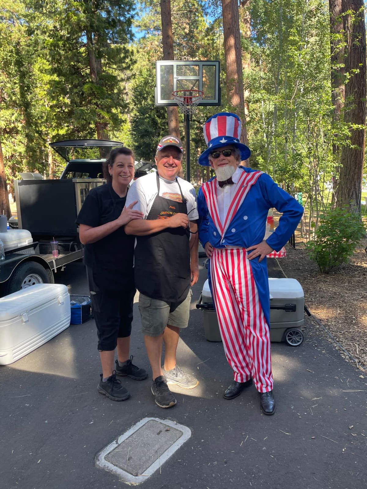 Two Carson City BBQ crew members in aprons posing beside a person costumed as Uncle Sam at an outdoor Fourth of July event, their cooker parked behind them.