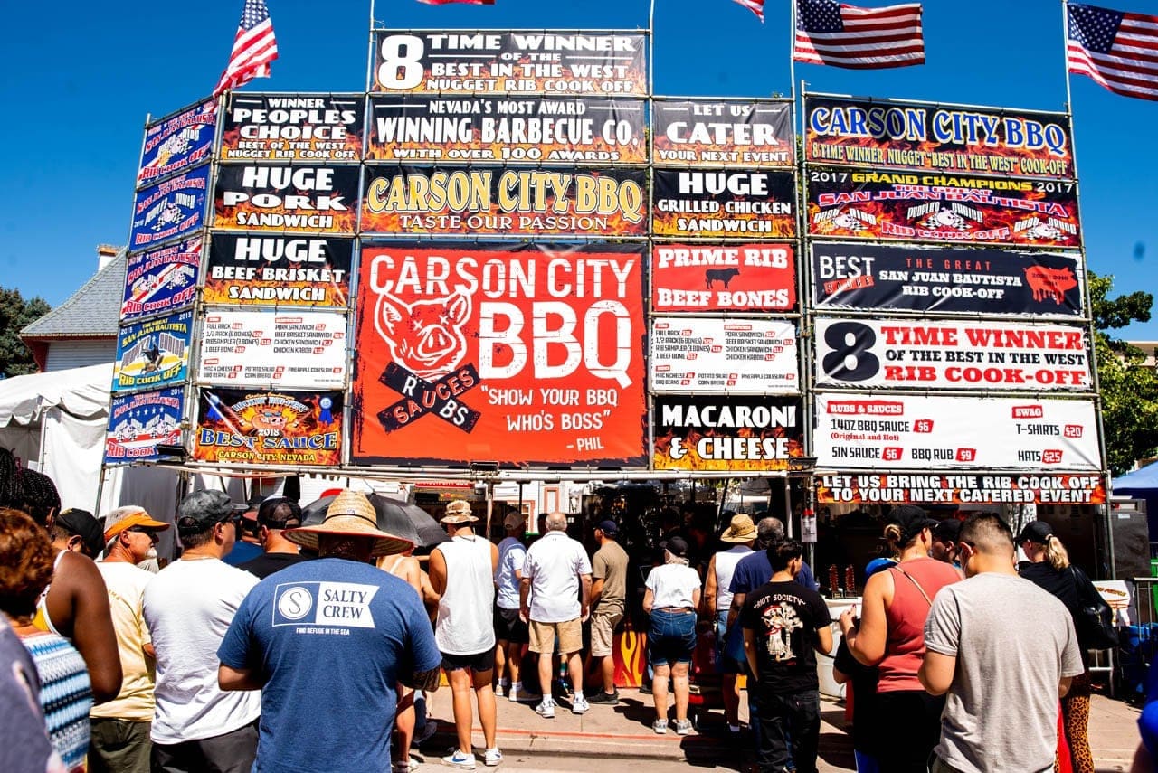 The full Carson City BBQ booth facade at a rib cook-off, its stacked signs topped by an "8 time winner of the Best in the West Rib Cook-Off" banner, with a queue of customers below.
