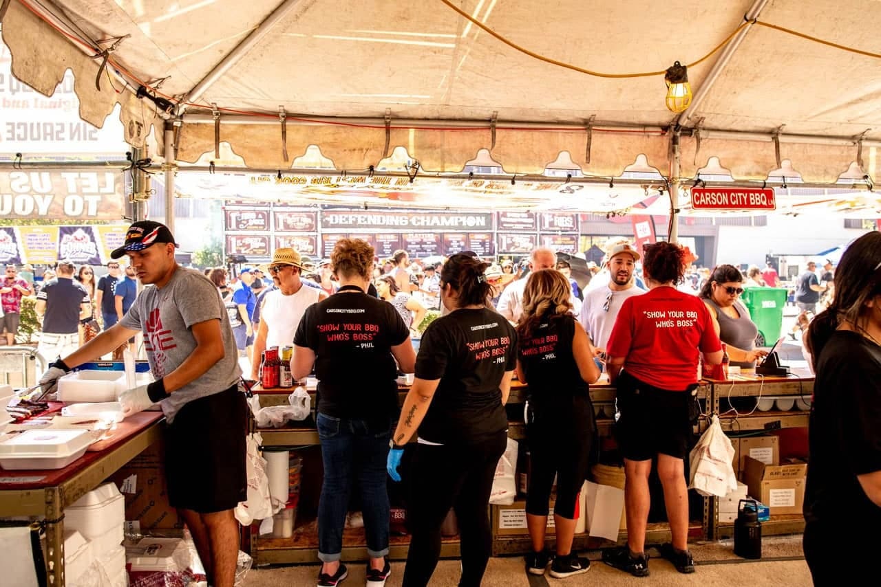 The Carson City BBQ crew working shoulder to shoulder along the service counter inside their tent, plating orders while the festival crowd passes outside.