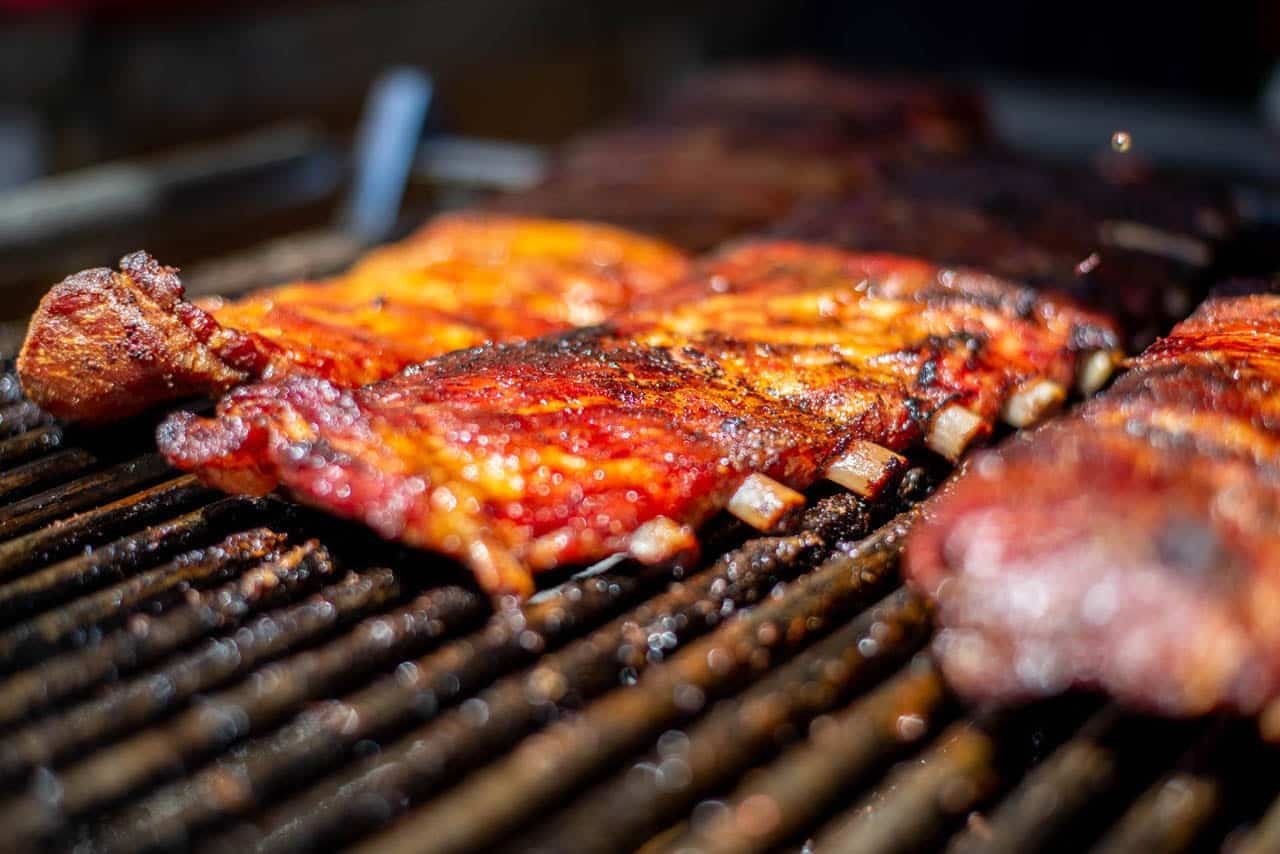 Whole racks of glossy, sauce-glazed ribs cooking across a wide charcoal grill.