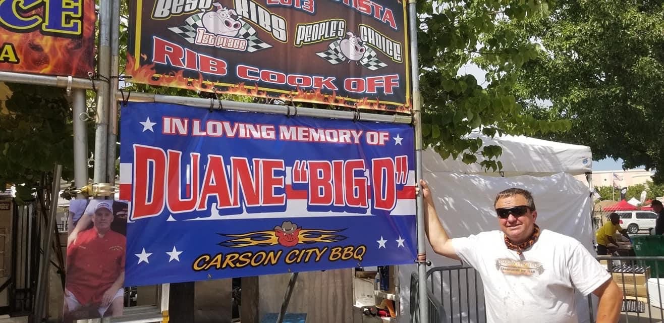 A memorial banner reading "In loving memory of Duane 'Big D'" hung on the Carson City BBQ cook-off booth, with a man standing beside it holding his hand to his heart.