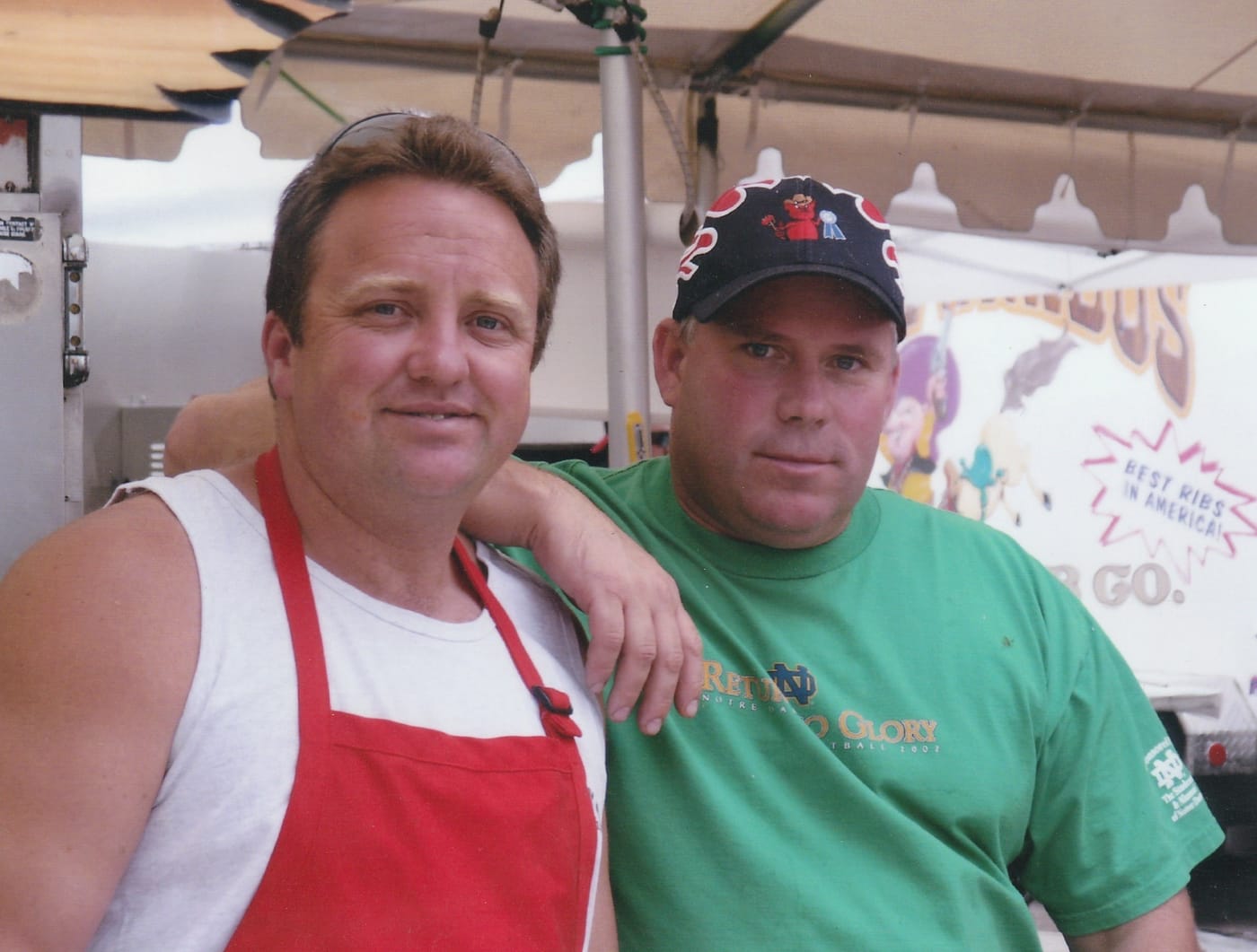 Carson City BBQ founders Phil Hyatt and Duane Felker side by side under a cook-off canopy, one in a red apron over a white tank top, the other in a green t-shirt and a ball cap.