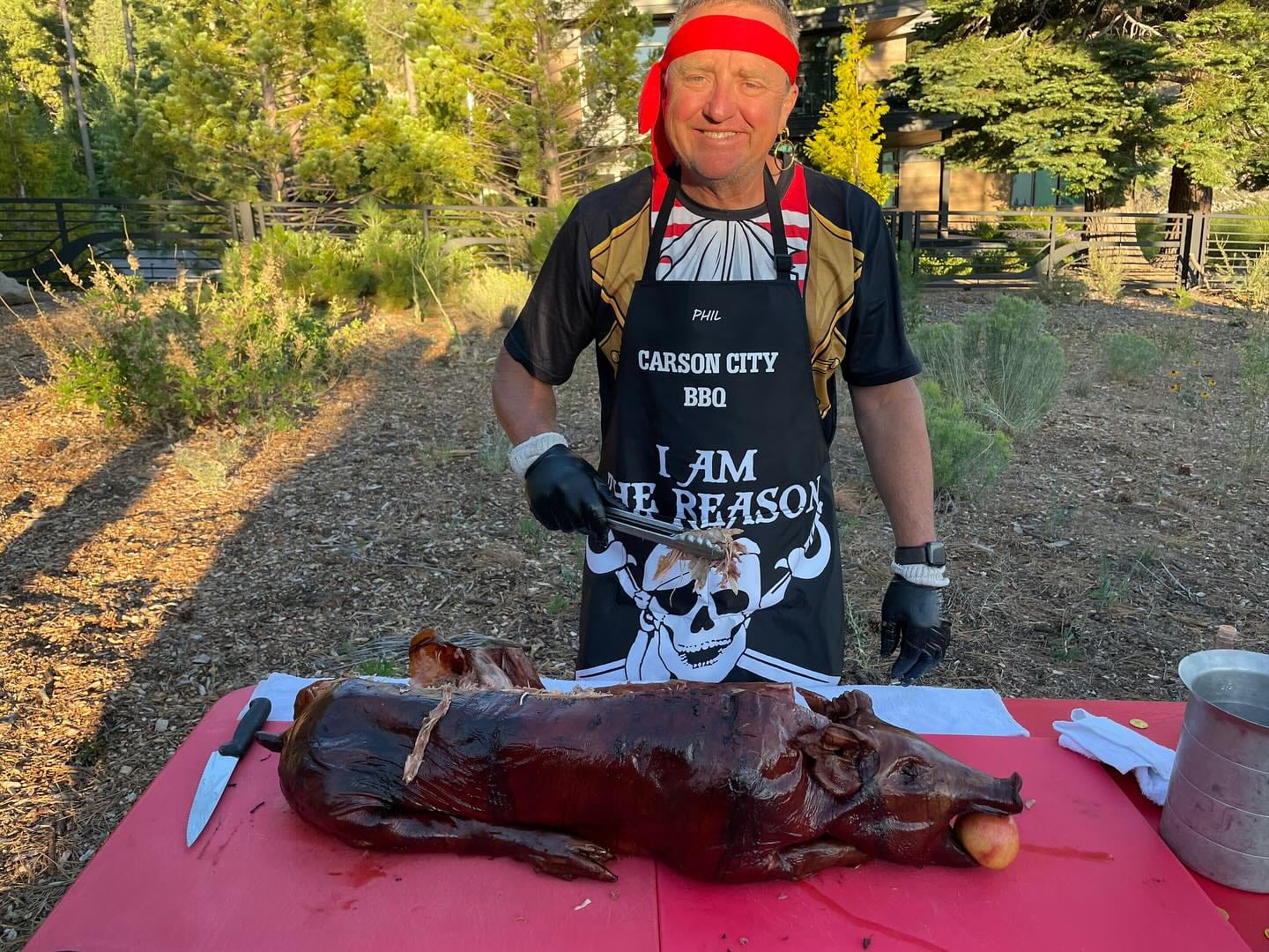 A cook in a Carson City BBQ apron and red bandana standing over a whole roasted hog on a red cutting board at an outdoor event.