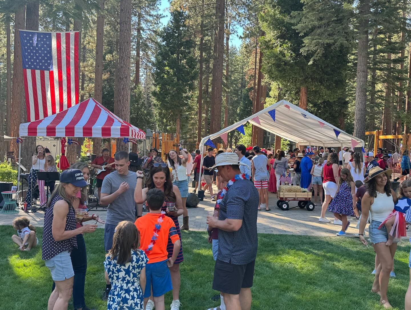 Guests gathered on the grass between an American-flag-print canopy and a white canopy at an outdoor event in a pine forest clearing.