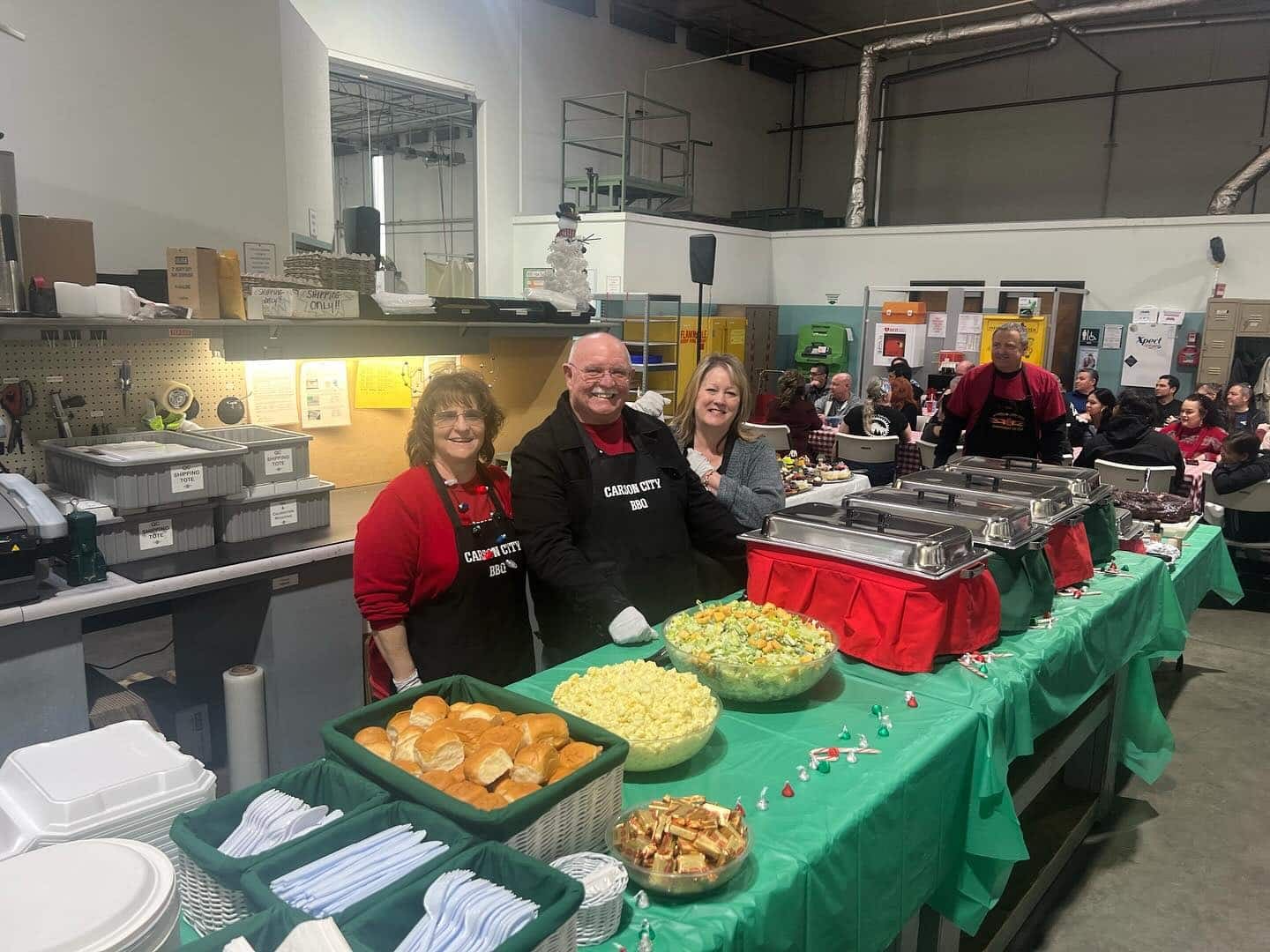 Three staff in Carson City BBQ aprons standing behind an indoor buffet line dressed with green tablecloths, serving trays of corn, rolls and salads.