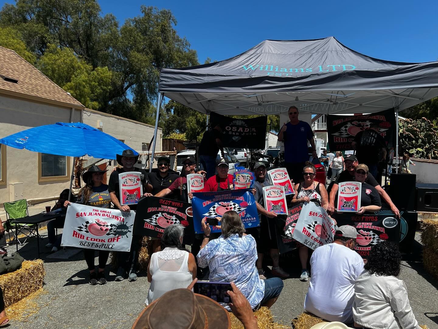 A crowd seated on hay bales under a canopy at a rib cook-off, many of them holding up printed fan signs for Carson City BBQ.