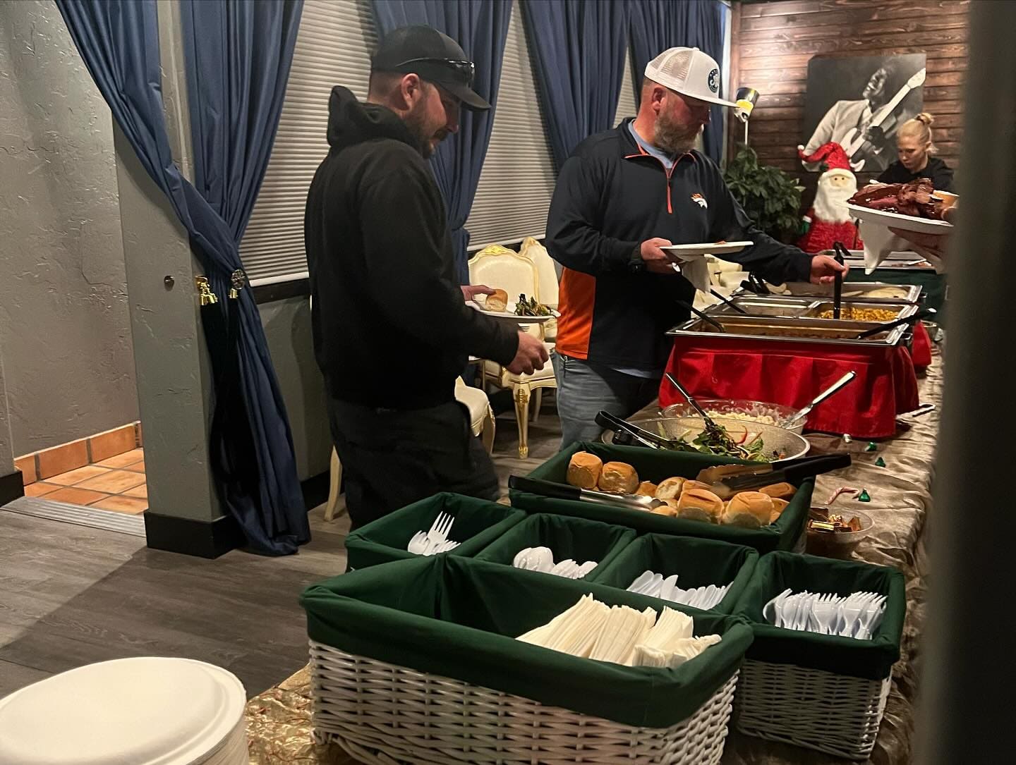 Guests serving themselves from a chafing-dish buffet along a draped wall inside a banquet room, with baskets of rolls and cutlery caddies at the end of the line.