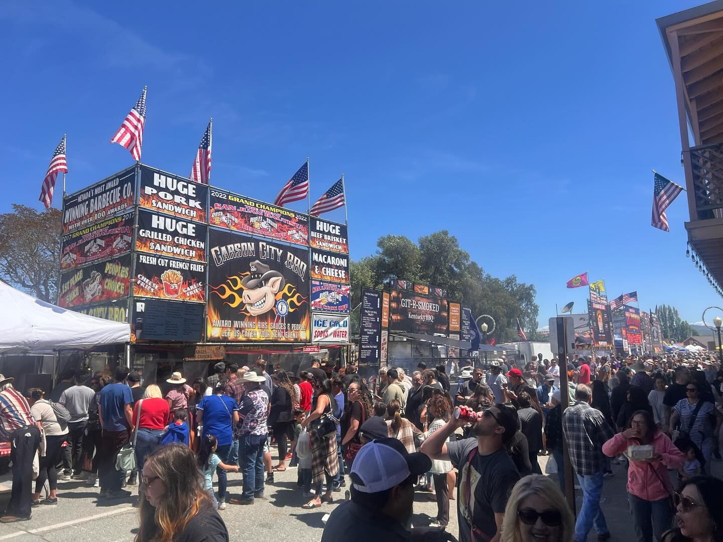 A dense crowd filling the midway at the Best in the West Nugget Rib Cook-Off, walking past the tall illuminated Carson City BBQ booth signs advertising ribs, macaroni and cheese and grilled chicken.
