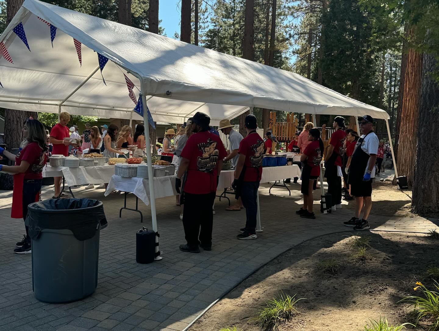 Staff in red shirts working a long buffet line under a white marquee strung with patriotic bunting at a forest event.
