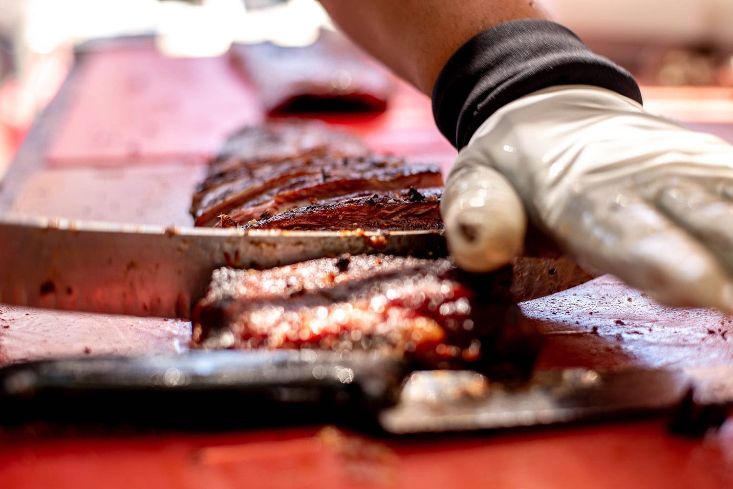 A gloved crew member portioning a rack of ribs on a red cutting board with a long knife during cook-off service.