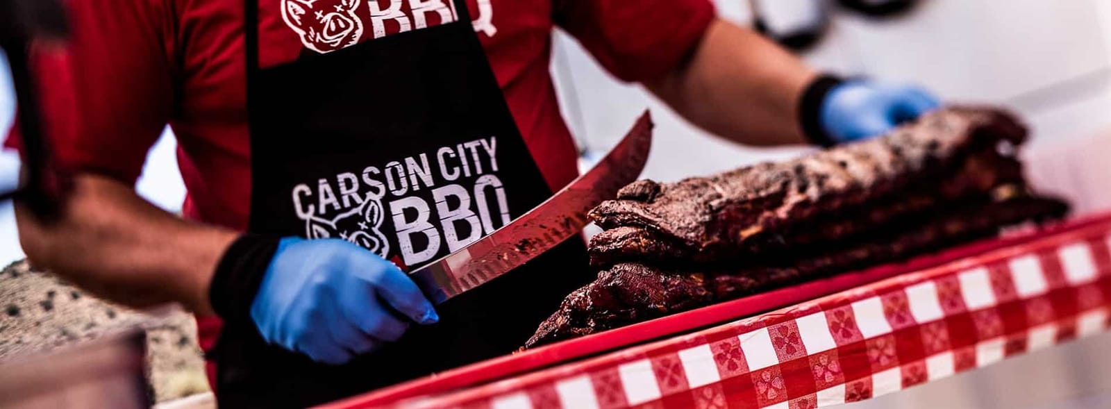 A cook in a Carson City BBQ apron slicing into a rack of ribs on a red-and-white checked cloth.