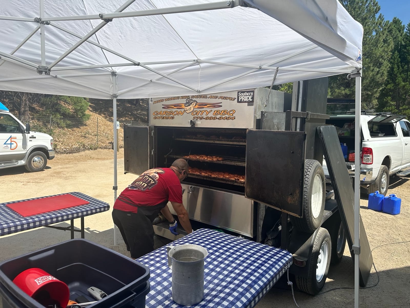 A crew member loading the Carson City BBQ rotisserie trailer beneath a pop-up canopy at an outdoor event site.