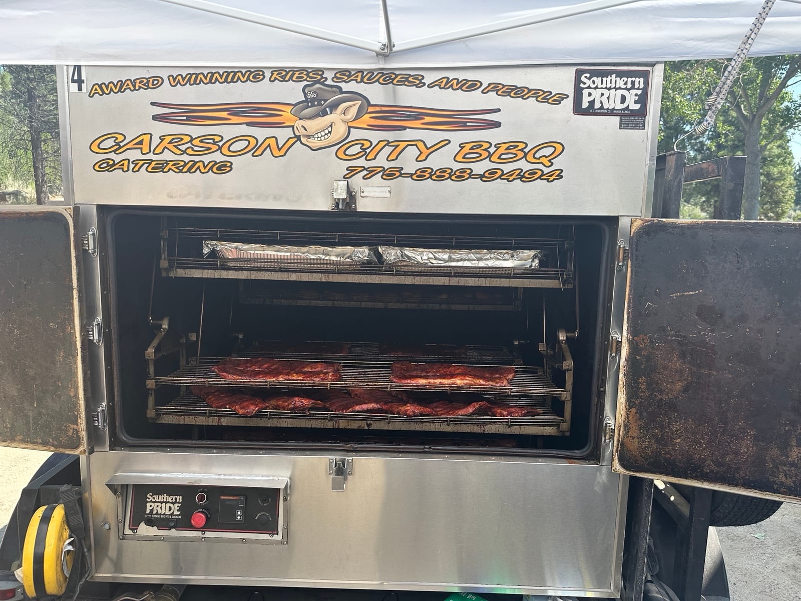 The Carson City BBQ Southern Pride rotisserie trailer with its doors open, racks of meat visible inside, the trailer signwritten "Award winning ribs, sauces and people".