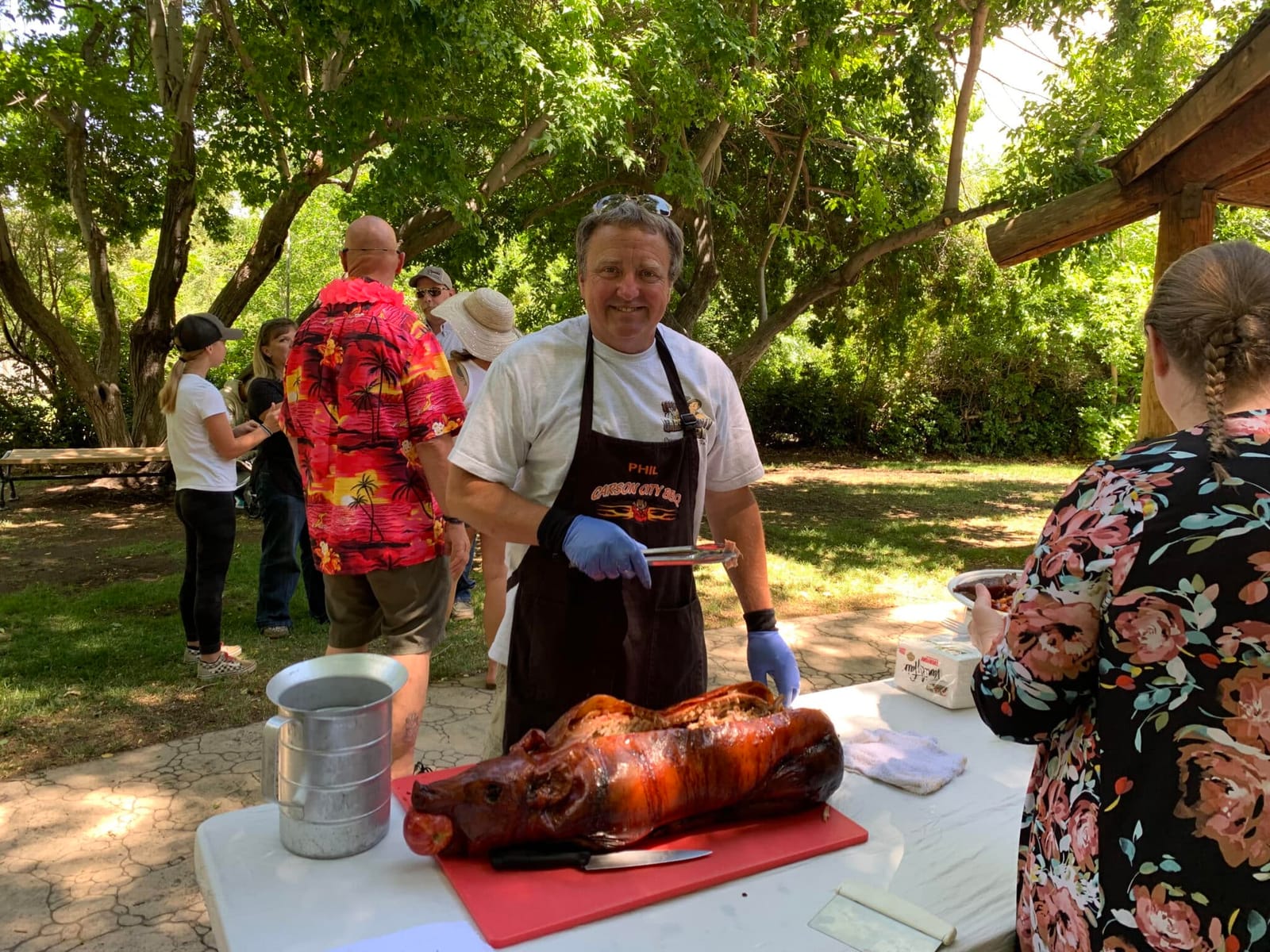 Guests gathered around a cook carving a whole roasted hog on a table under the trees at a garden event.