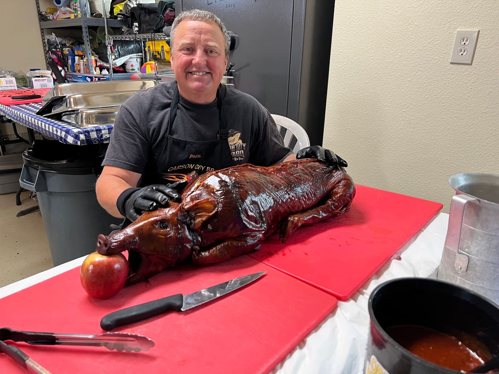 A cook in a Carson City BBQ apron smiling behind a whole roasted suckling pig laid out on a red cutting board, carving knife and tongs beside it.