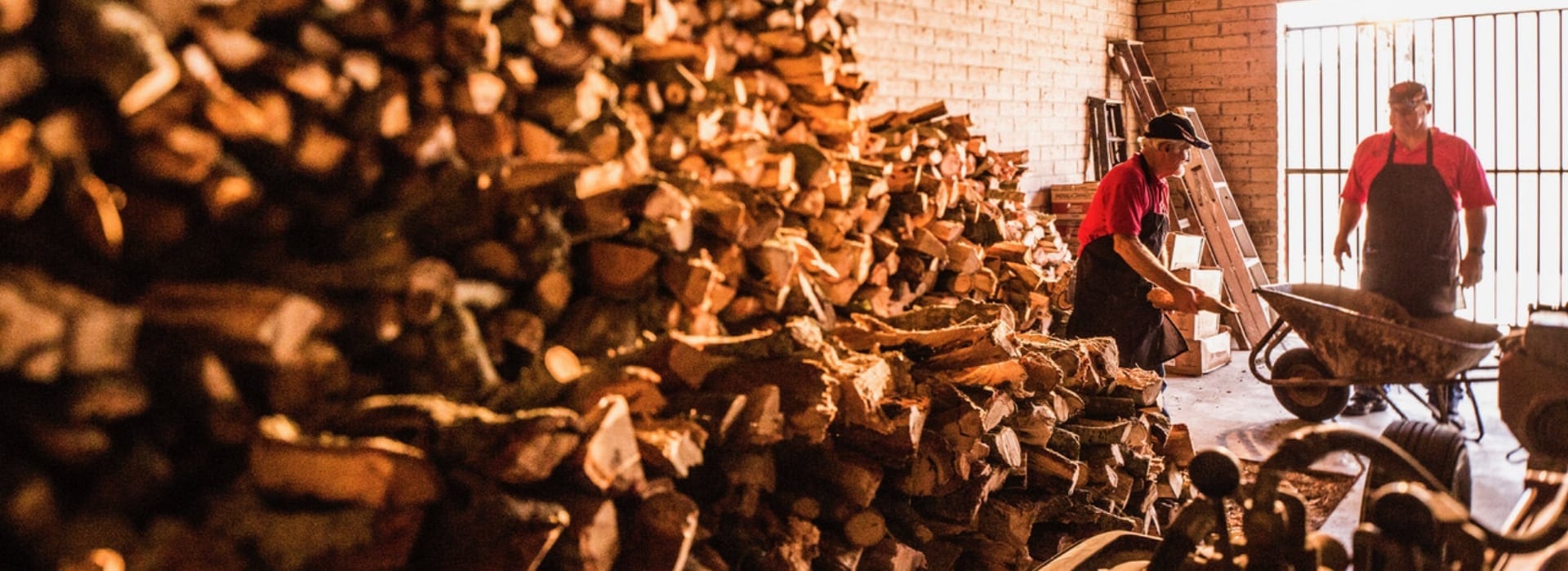 Two Carson City BBQ cooks in branded aprons loading split firewood into a wheelbarrow beside the stacked woodpile in their shed.