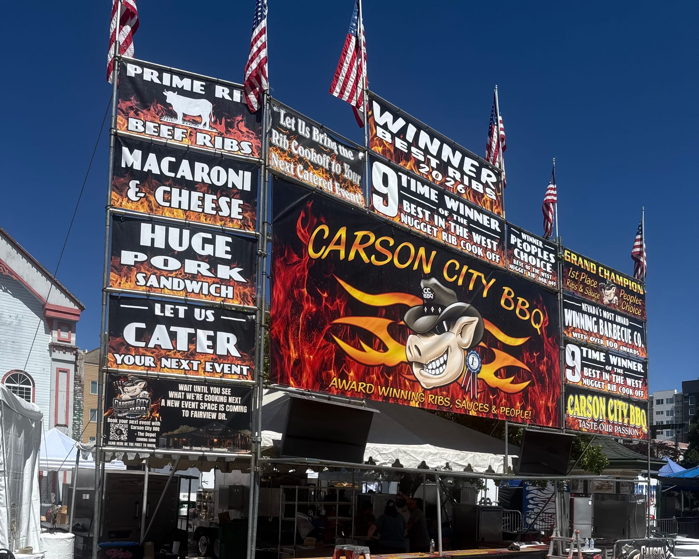 The Carson City BBQ cook-off booth under a blue sky, American flags flying above a wall of banners. The centrepiece is a flaming “Carson City BBQ” sign with the pig mascot and “Award winning ribs, sauces & people!”. Boards around it read “Winner Best Ribs 2026”, “9 time winner Best in the West Nugget Rib Cook Off”, “Peoples Choice” and “Grand Champion”, beside menu boards for prime rib and beef ribs, macaroni & cheese, a huge pork sandwich and “Let us cater your next event”.
