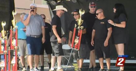 Cook-off competitors gathered on stage behind their trophies as a television camera crew films the awards, a Channel 2 microphone flag in the foreground.