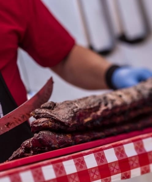 A gloved cook portioning racks of dark-crusted pork ribs with a long knife on a red-and-white checked cloth.