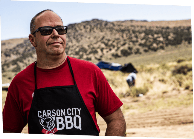 Devin Stone in sunglasses, a red shirt and a black Carson City BBQ apron, standing outdoors on open Nevada high desert with hills in the background.