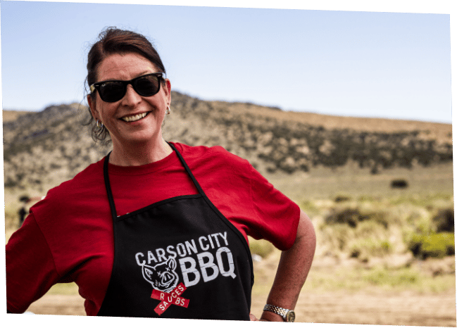 Michele Wilkinson smiling in sunglasses, a red shirt and a black Carson City BBQ apron, standing outdoors with dry Nevada hills and sagebrush behind her.