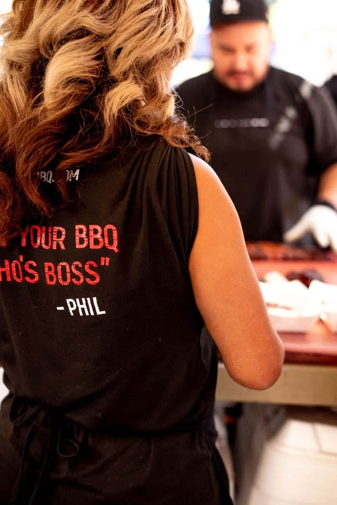 A crew member seen from behind in a black Carson City BBQ apron printed "Show your BBQ who's boss — Phil", working the prep table alongside a colleague.