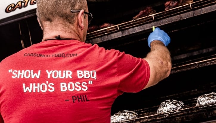 The back of a red Carson City BBQ shirt reading "Show your BBQ who's boss — Phil", worn by a cook reaching into the smoker to tend the racks.