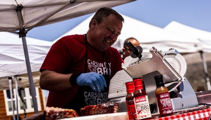 Phil Hyatt working a meat slicer at an outdoor catering station under a canopy, bottles of Carson City BBQ sauce and a tray of ribs on the table beside him.
