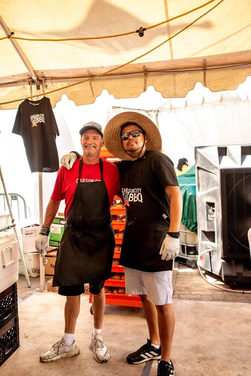 Two Carson City BBQ crew members in black aprons, one in a straw hat, standing arm in arm inside the team's tent beside a rack of merchandise shirts.