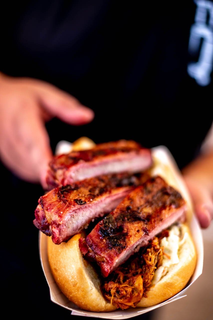 Hands passing a paper tray holding a rib sandwich — sauced ribs stacked on a soft bun — toward the camera.