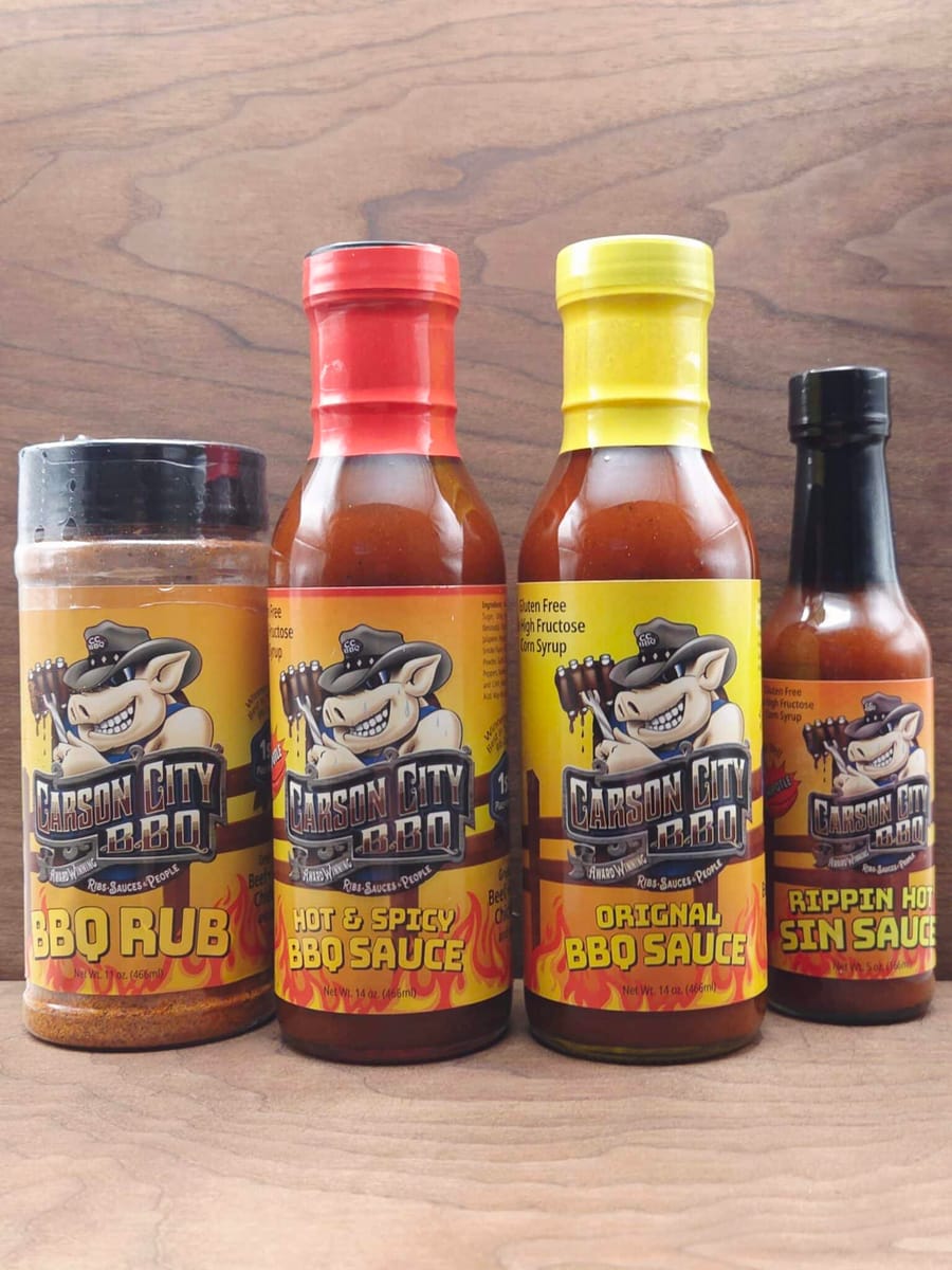 All four Carson City BBQ products on a wooden table: the BBQ Rub shaker, Hot & Spicy BBQ Sauce, Original BBQ Sauce and Chipotle Sin Sauce