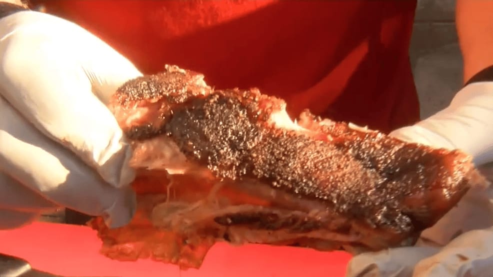 A close-up filmed for a television segment: gloved hands pulling apart a rack of ribs to show the smoke ring, against a red background.
